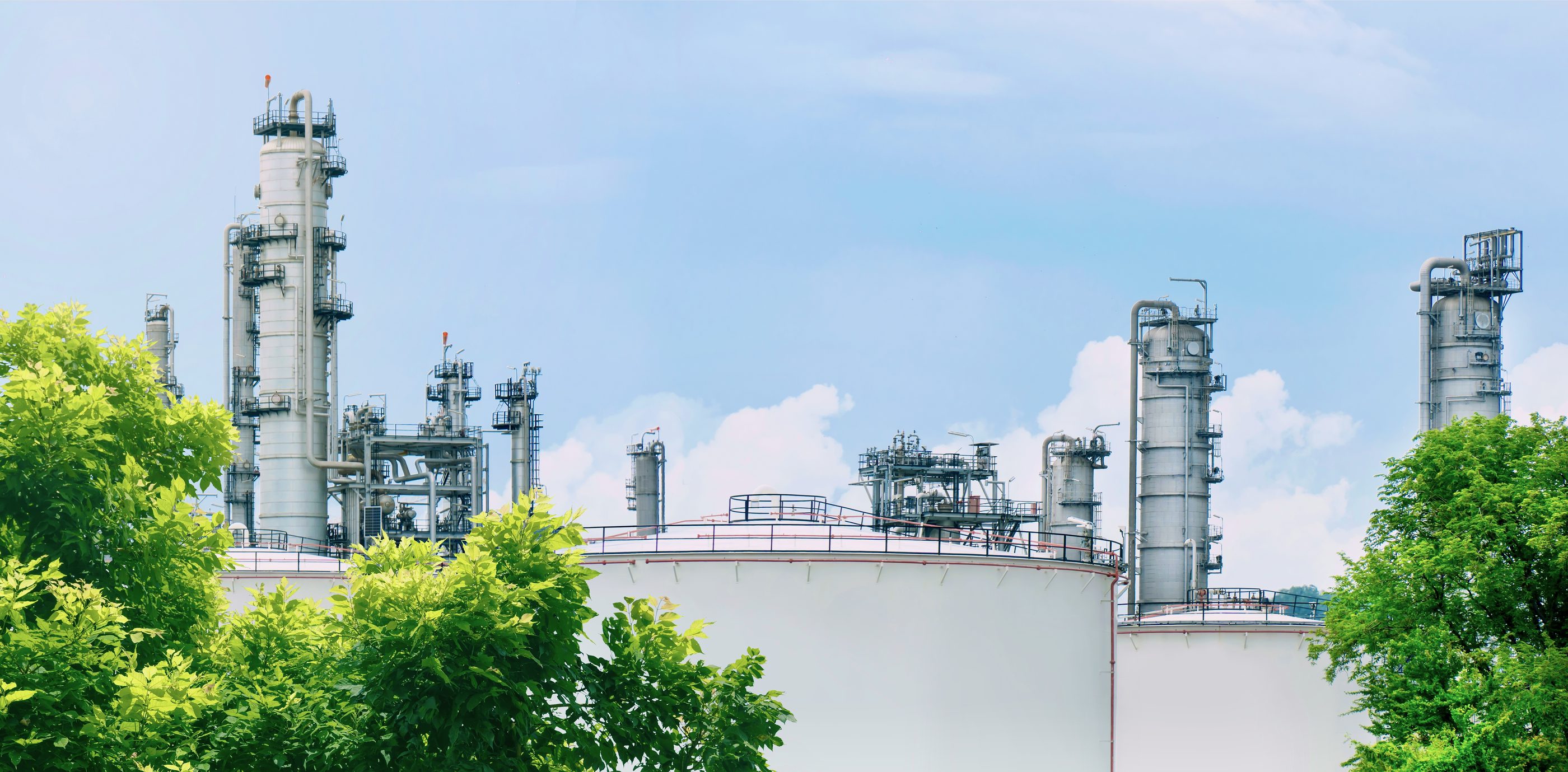 White storage tanks and process towers rising above green trees under a summer sky